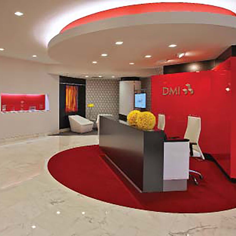 A receptionist desk in an office surrounded by a modern workspace design that features a red back wall and rug.